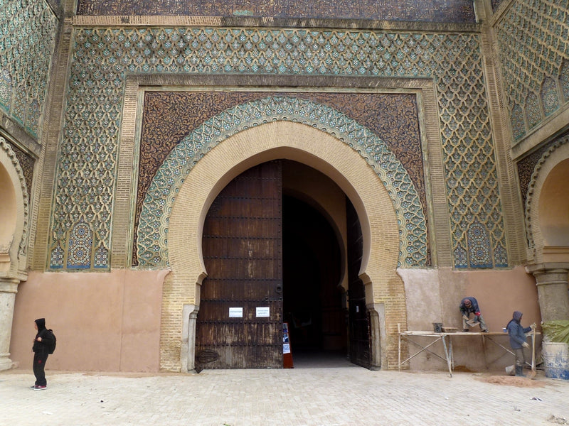 Bab Mansour in Meknès, with its decorated horseshoe arch, partly open wooden doors, pedestrians and workers beside the entrance.