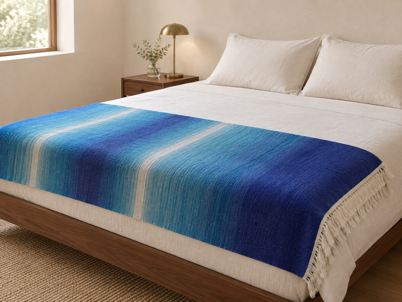 Illustrative styling scene with the Blue Sky blanket's blue, aqua and white bands across the foot of an ivory bed in a warm contemporary bedroom.