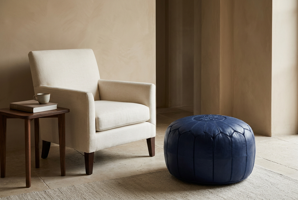 Dark blue Moroccan leather pouf with stitched panels beside an ivory armchair in a warm interior.