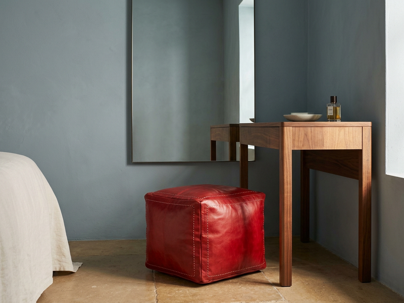 Salwa Red square leather pouf beside a walnut dressing table, with a blue-grey wall and ivory bedding.