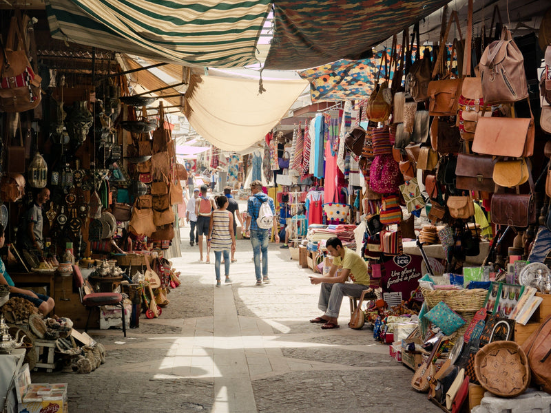 Handmade Moroccan leather bags hanging in a Marrakesh medina souk alleyway — cognac, tan and terracotta tones, traditional craft