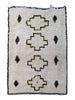 A rectangular woven rug with a cream-colored pile, featuring four large central stepped motifs and six small diamond motifs, outlined in olive green and finished with a black fringed border.