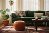 A cozy living room scene featuring a deep green velvet sofa with floral throw pillows, a round brown leather pouf, a dark wood coffee table with a lit candle, and a patterned area rug on a light wood floor.