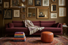 A styled living room scene featuring a deep burgundy velvet sofa as the main product, decorated with a cream throw blanket. The sofa is set against a dark brown wall covered in framed art, with a leather pouf, a stack of books, and a patterned rug in the foreground.